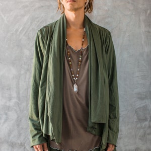 May include: A person wearing a green long-sleeved jacket with a brown shirt underneath. The jacket has a shawl collar and is open at the front. The person is wearing a beaded necklace with a silver pendant. The person has dreadlocks and is wearing a white headband.