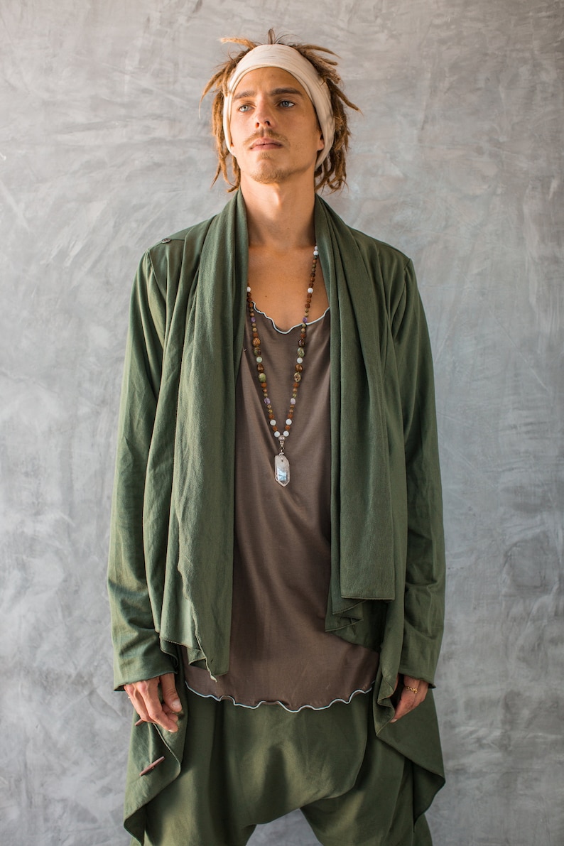 May include: A person wearing a green long-sleeved jacket with a brown shirt underneath. The jacket has a shawl collar and is open at the front. The person is wearing a beaded necklace with a silver pendant. The person has dreadlocks and is wearing a white headband.