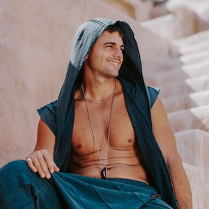 May include: A person wearing a teal hooded vest and matching pants. A silver necklace with a dark pendant is visible. The person is sitting on a light-colored stone structure, with a soft, natural light.