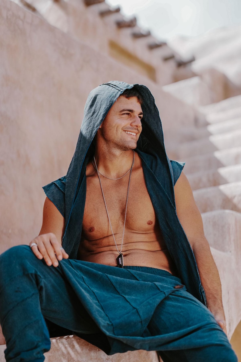 May include: A person wearing a teal hooded vest and matching pants. A silver necklace with a dark pendant is visible. The person is sitting on a light-colored stone structure, with a soft, natural light.