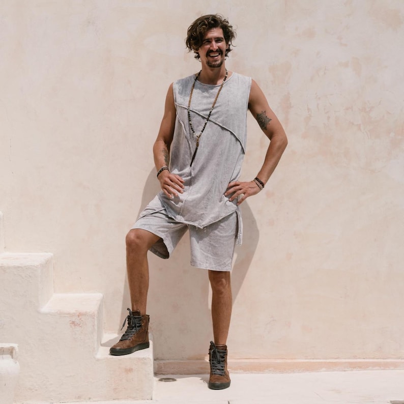 May include: A man wearing a grey sleeveless shirt with a beaded necklace and grey shorts. He is standing on a set of stairs with a white wall behind him. He is wearing brown leather boots.