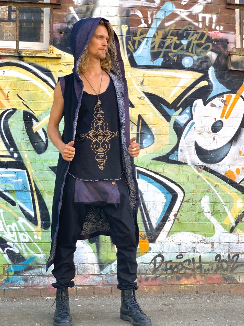 May include: A black sleeveless top with a gold geometric design, paired with black pants and a long hooded vest. The vest has a dark blue hood and a patterned lining. The outfit is completed with black boots. The background features colorful graffiti art.