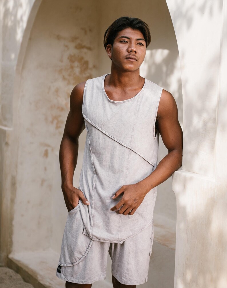 May include: A person wearing a light grey sleeveless shirt with a diagonal seam detail and matching light grey shorts. The person is standing in a doorway with a white wall behind them.