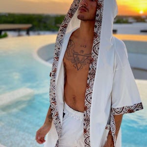 May include: A white robe with brown and blue patterned trim. The robe has a hood and is open in the front, revealing a person's bare chest with a tattoo. The person is standing near a pool.