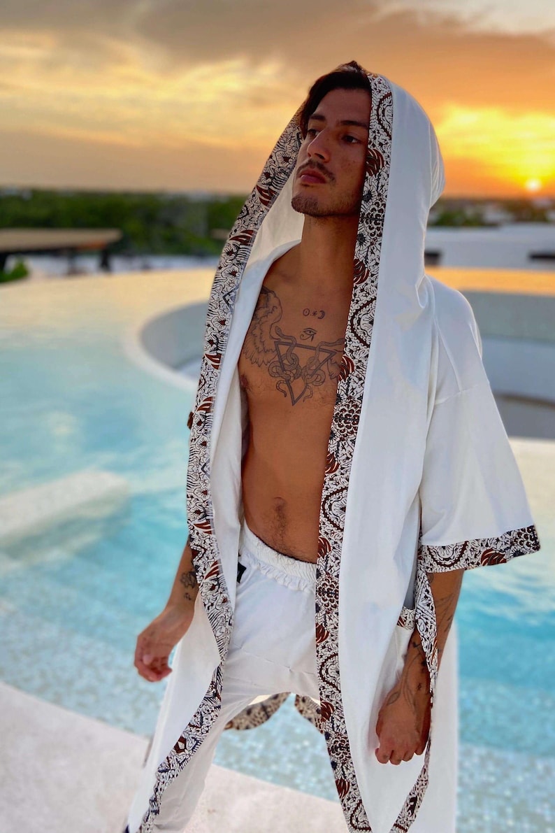May include: A white robe with brown and blue patterned trim. The robe has a hood and is open in the front, revealing a person's bare chest with a tattoo. The person is standing near a pool.