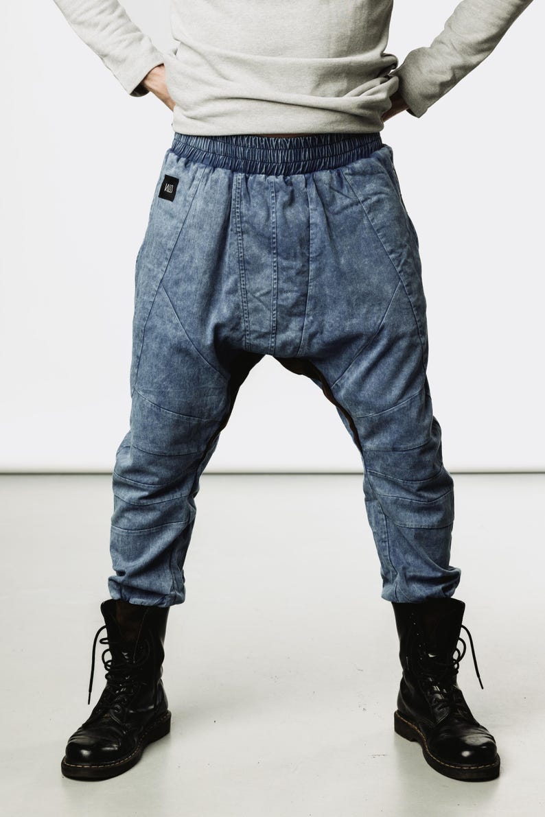 May include: Blue denim-style pants with a unique, relaxed fit. The pants feature a gathered waistband and stitched details. Paired with black combat boots, the pants have a distinctive, modern style. The model is wearing a gray long-sleeve shirt.