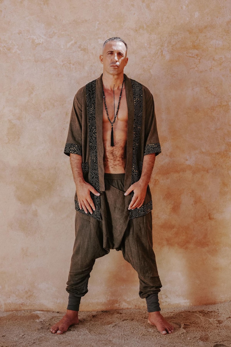 May include: A brown linen kimono-style jacket with black geometric trim, paired with matching harem pants. A long beaded necklace with a tassel hangs down. The outfit is a relaxed, bohemian style.