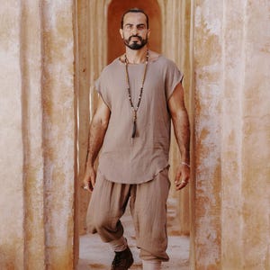 May include: A man wearing a light brown linen outfit, including a short-sleeved top and cropped pants. He is also wearing a long beaded necklace and brown boots. The background features a series of arched doorways.