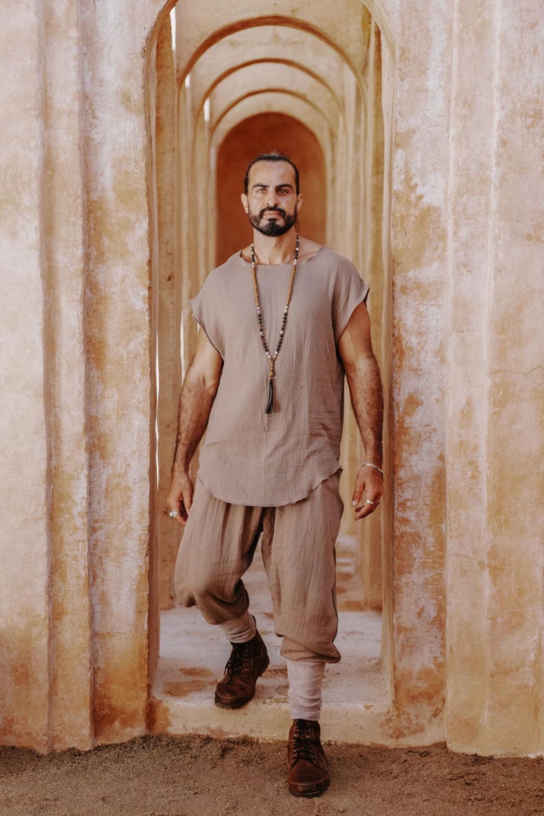 May include: A man wearing a light brown linen outfit, including a short-sleeved top and cropped pants. He is also wearing a long beaded necklace and brown boots. The background features a series of arched doorways.