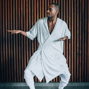 May include: A person wearing a white robe with a brown beaded necklace, standing in front of a wood panel wall. The person is looking to the right and has their arms outstretched.