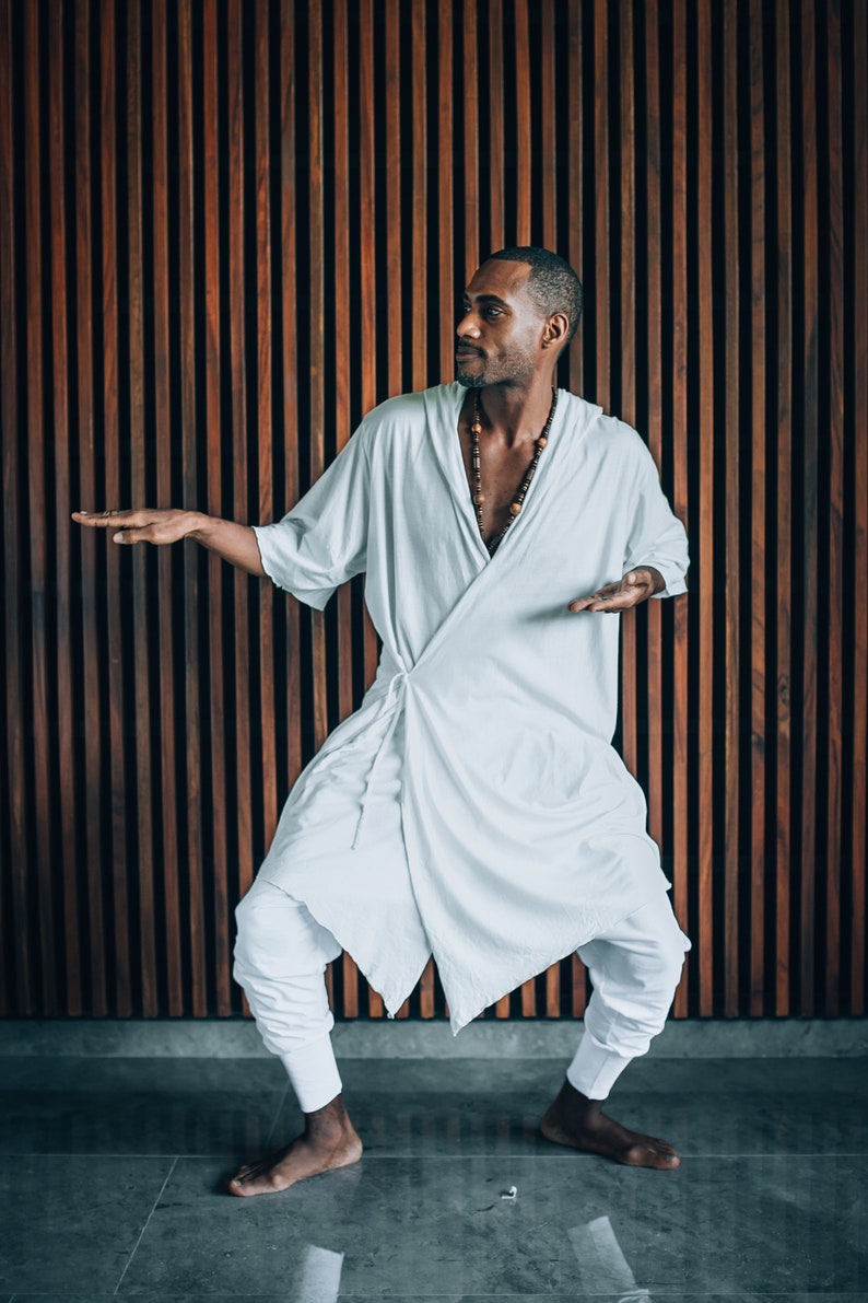 May include: A person wearing a white robe with a brown beaded necklace, standing in front of a wood panel wall. The person is looking to the right and has their arms outstretched.