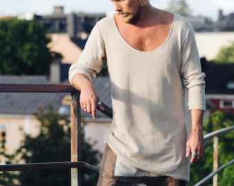 May include: A long-sleeved, beige, ribbed knit shirt with a wide, rounded scoop neckline. The shirt features a straight hemline and a lightweight, textured fabric construction that falls to the mid-thigh area, worn over dark brown trousers.