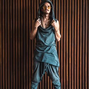 May include: A person wearing a dark green hooded top with a long, flowing design. They are wearing round, yellow-tinted sunglasses and several necklaces. The person is standing in front of a wall of vertical wooden slats.