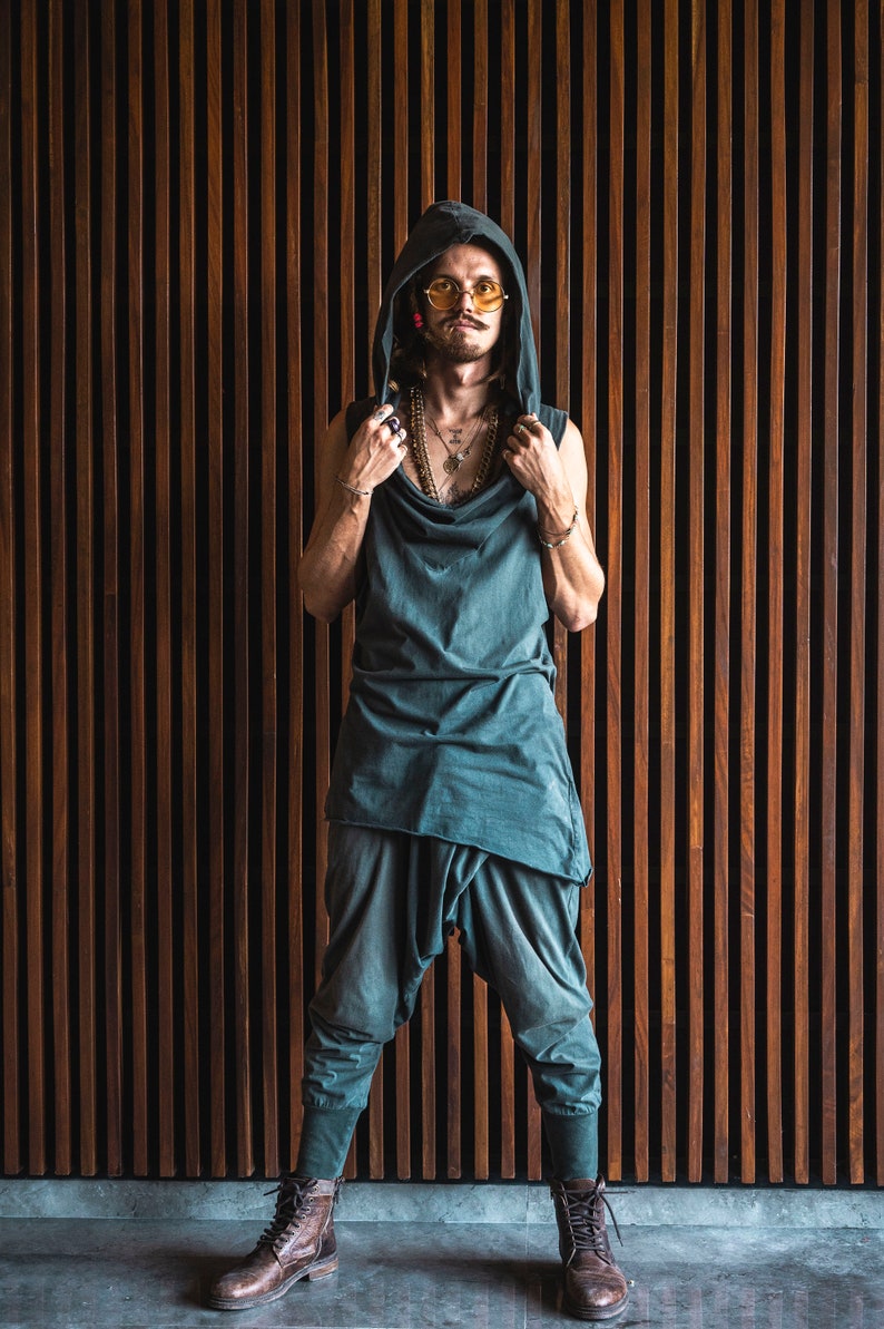 May include: A person wearing a dark green hooded top with a long, flowing design. They are wearing round, yellow-tinted sunglasses and several necklaces. The person is standing in front of a wall of vertical wooden slats.