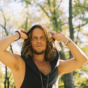May include: A man with long, curly hair is wearing a black hooded sleeveless shirt. He is standing in a forest with trees in the background.