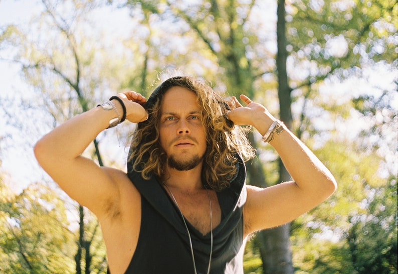May include: A man with long, curly hair is wearing a black hooded sleeveless shirt. He is standing in a forest with trees in the background.