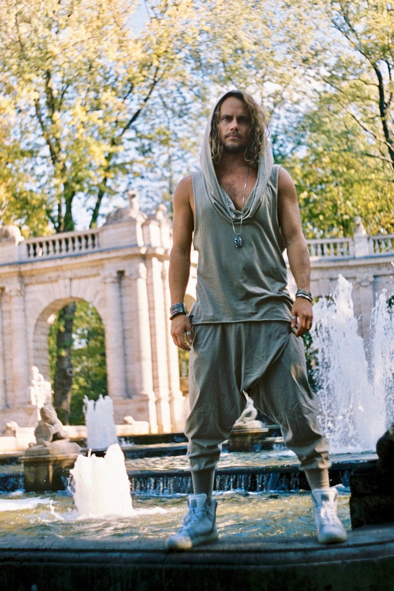 May include: A man wearing a gray hooded top with a silver necklace stands in front of a fountain. He is wearing gray pants and white sneakers.