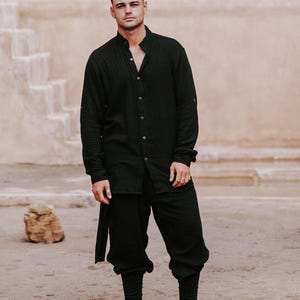 May include: A person wearing a black long-sleeve shirt with a mandarin collar and matching black pants. The outfit includes black boots. The background features a beige wall and a sandy ground.