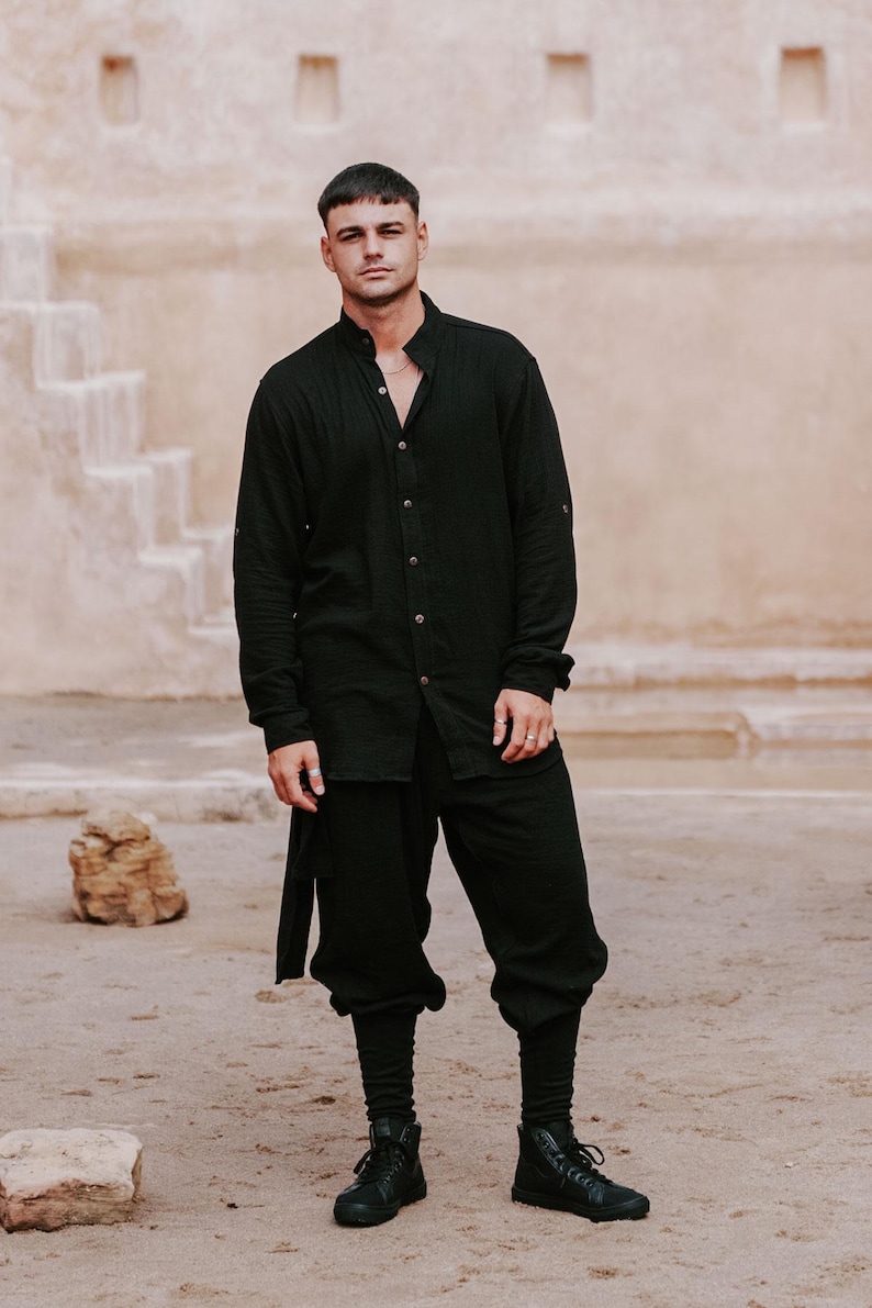May include: A person wearing a black long-sleeve shirt with a mandarin collar and matching black pants. The outfit includes black boots. The background features a beige wall and a sandy ground.