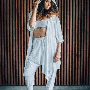 May include: A woman wearing a white hooded robe over a white crop top and white trousers. The robe has a long, flowing design and the trousers are loose-fitting. She is standing in front of a wooden wall.