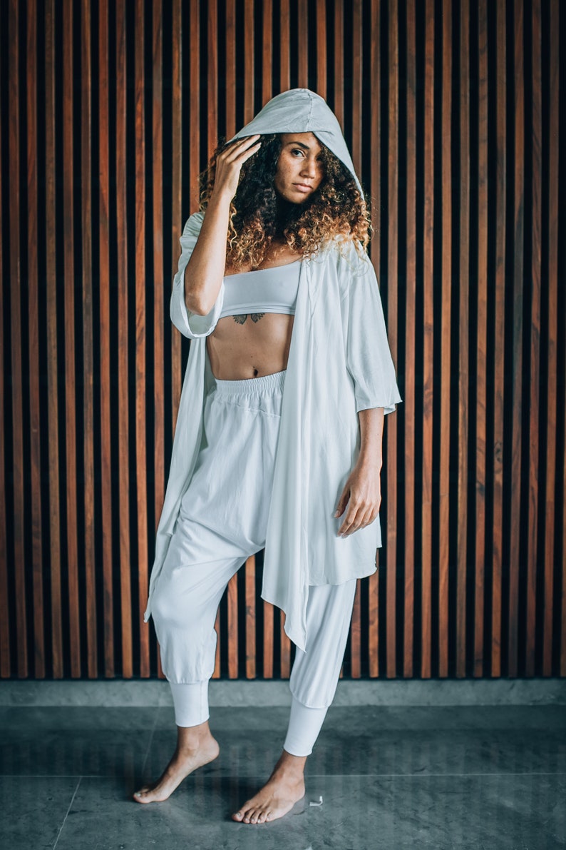 May include: A woman wearing a white hooded robe over a white crop top and white trousers. The robe has a long, flowing design and the trousers are loose-fitting. She is standing in front of a wooden wall.