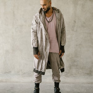 May include: A person wearing a light brown hooded coat, a pink shirt, and matching pants. The coat has black trim and the person is wearing black high-top sneakers. The outfit is a neutral color.