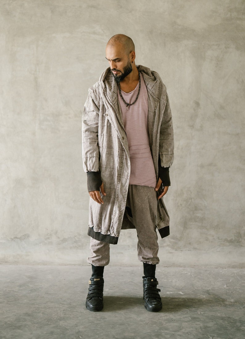May include: A person wearing a light brown hooded coat, a pink shirt, and matching pants. The coat has black trim and the person is wearing black high-top sneakers. The outfit is a neutral color.