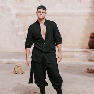 May include: Black linen shirt and pants ensemble. The long-sleeved shirt is open at the chest, and the pants are a loose, cropped style. The outfit is paired with black boots. The background features a beige building and sandy ground.