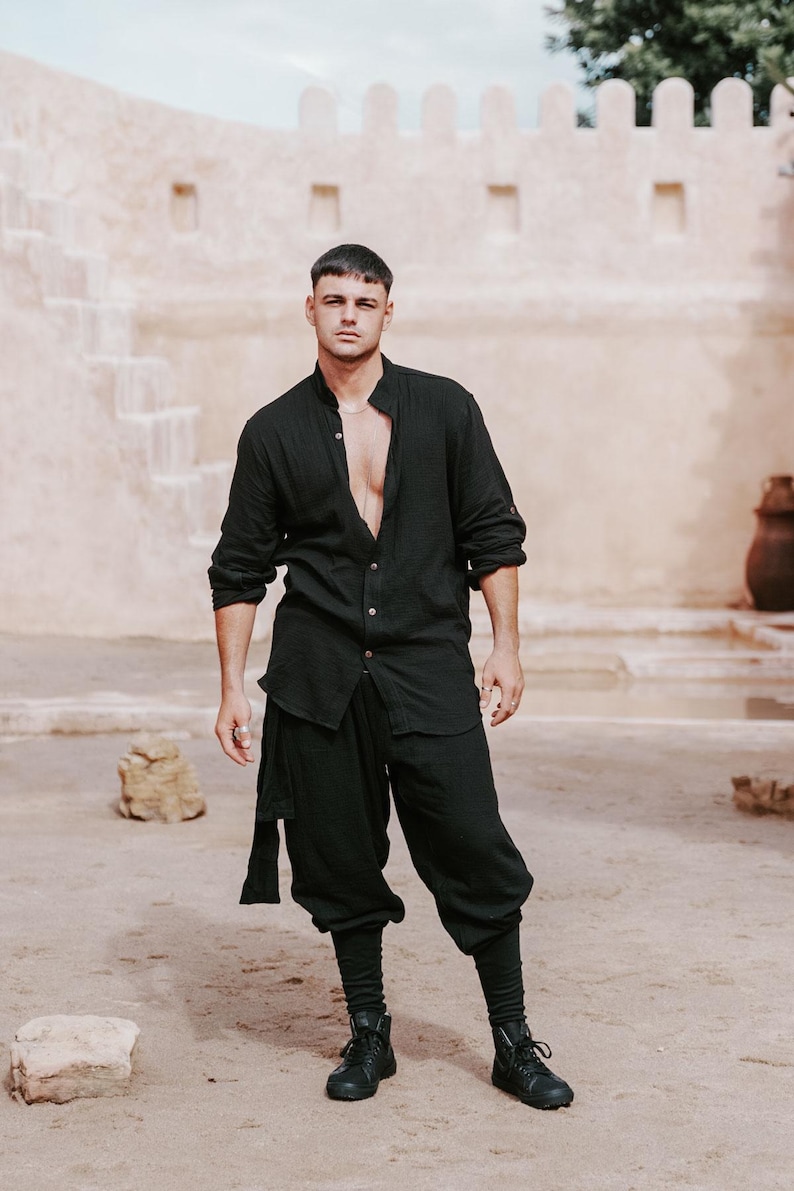 May include: Black linen shirt and pants ensemble. The long-sleeved shirt is open at the chest, and the pants are a loose, cropped style. The outfit is paired with black boots. The background features a beige building and sandy ground.