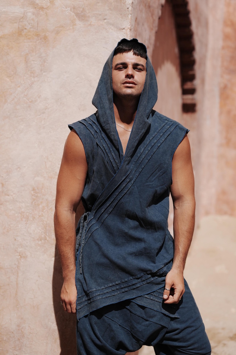 May include: A sleeveless, hooded wrap top and matching pants in a dark blue denim-like fabric. The top features pleats and a wrap design with a tie closure. The model is standing in front of a textured, peach-colored wall.