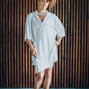 May include: A woman with curly brown hair is wearing a white, loose-fitting, wrap dress with a white shell necklace. She is standing in front of a wood panel wall and wearing black ankle boots.