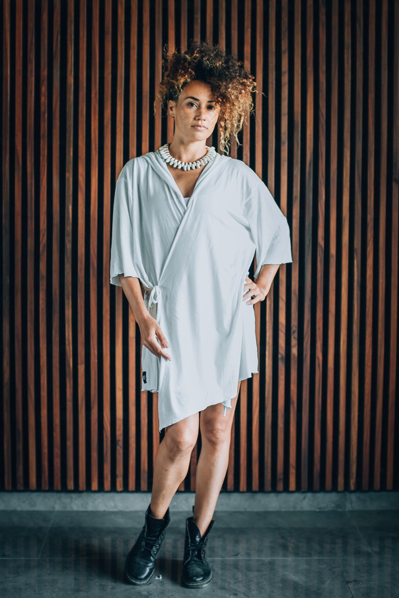 May include: A woman with curly brown hair is wearing a white, loose-fitting, wrap dress with a white shell necklace. She is standing in front of a wood panel wall and wearing black ankle boots.