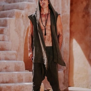 May include: A person wearing a dark olive green hooded vest, matching harem pants, and a long beaded necklace. The outfit is worn in a desert-like environment with stone steps and archways in the background.