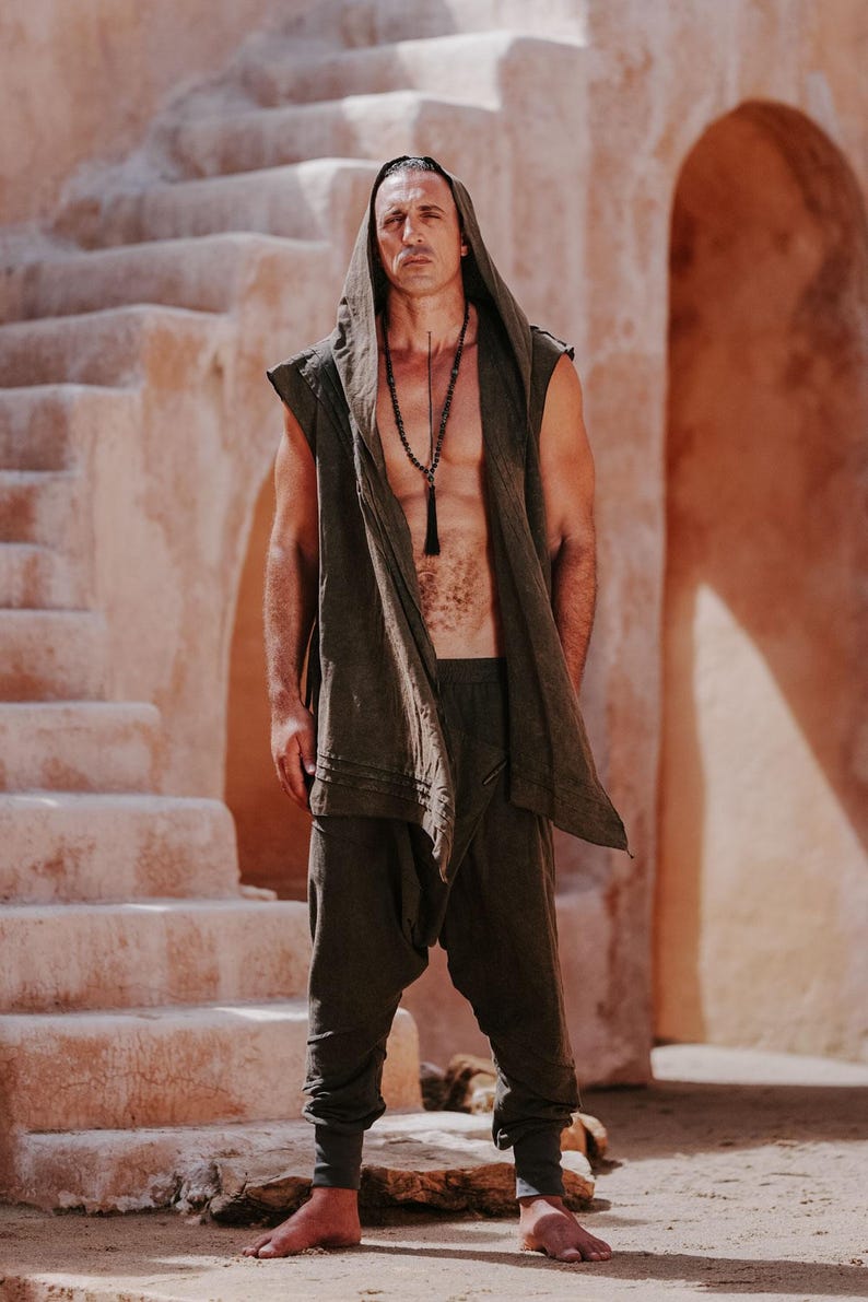 May include: A person wearing a dark olive green hooded vest, matching harem pants, and a long beaded necklace. The outfit is worn in a desert-like environment with stone steps and archways in the background.