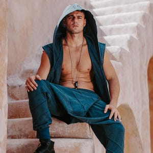 May include: A teal hooded vest and matching pants set. The vest has a sleeveless design and a hood. The pants have a relaxed fit. The model is wearing black high-top sneakers. The background is a light-colored stone structure.