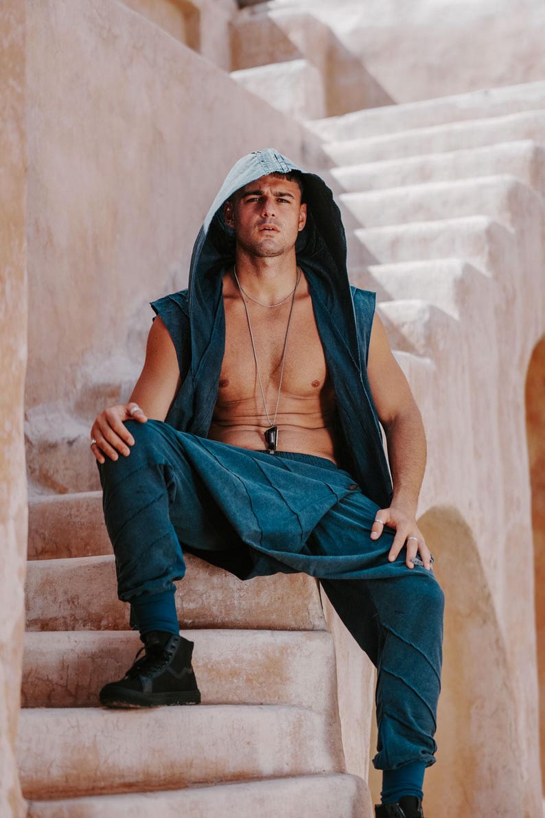 May include: A teal hooded vest and matching pants set. The vest has a sleeveless design and a hood. The pants have a relaxed fit. The model is wearing black high-top sneakers. The background is a light-colored stone structure.