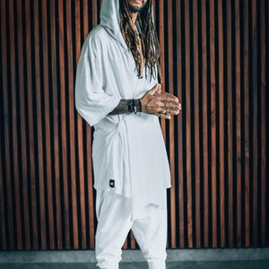 May include: A person wearing a white hooded robe with long dreadlocks. The person is standing in front of a wooden wall.