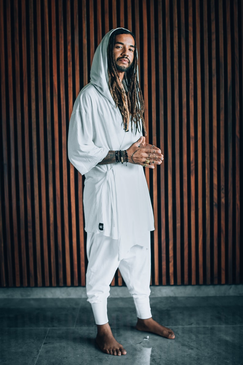 May include: A person wearing a white hooded robe with long dreadlocks. The person is standing in front of a wooden wall.
