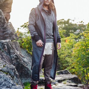 May include: A person wearing a gray hooded coat with purple trim, a gray sweater, and brown pants. The outfit includes a matching gray beanie and brown boots with red laces. The person is standing on a rocky surface.