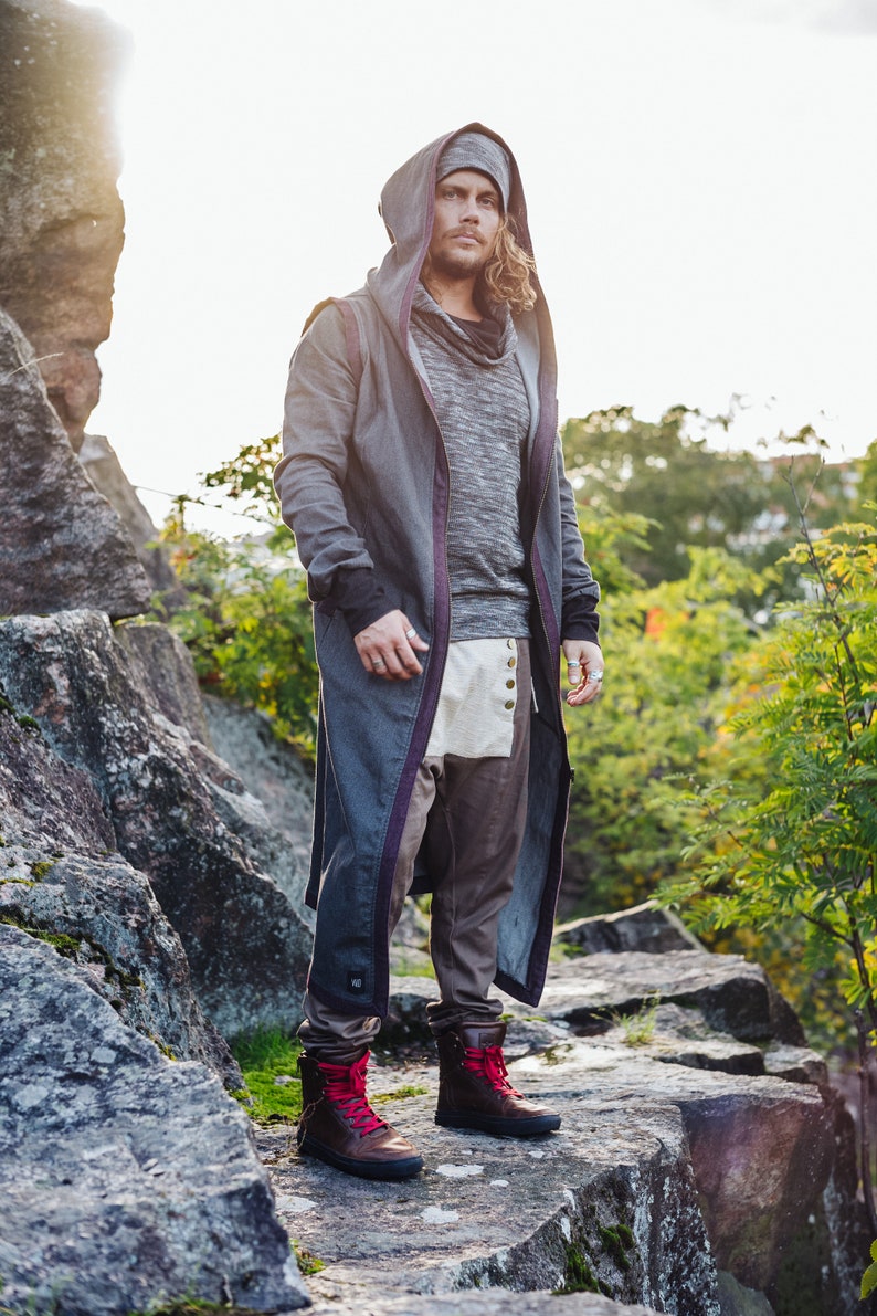May include: A person wearing a gray hooded coat with purple trim, a gray sweater, and brown pants. The outfit includes a matching gray beanie and brown boots with red laces. The person is standing on a rocky surface.