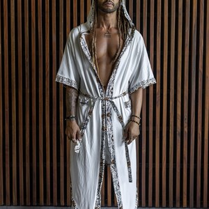 May include: A white robe with a brown and white patterned trim. The robe has a hood and is tied at the waist. The person wearing the robe has long dreadlocks and is standing in front of a wooden wall.
