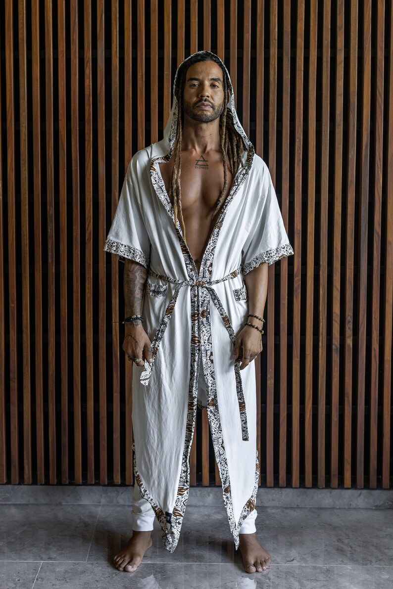 May include: A white robe with a brown and white patterned trim. The robe has a hood and is tied at the waist. The person wearing the robe has long dreadlocks and is standing in front of a wooden wall.