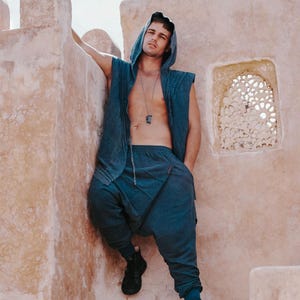 May include: A blue hooded vest and matching drop-crotch pants. The outfit is made of a soft fabric and is paired with black sneakers. The model is leaning against a textured, light-colored wall.