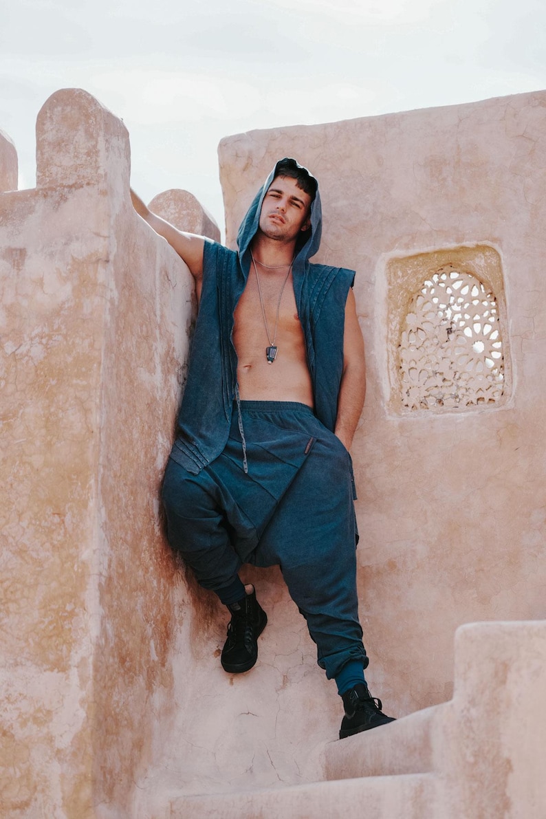 May include: A blue hooded vest and matching drop-crotch pants. The outfit is made of a soft fabric and is paired with black sneakers. The model is leaning against a textured, light-colored wall.