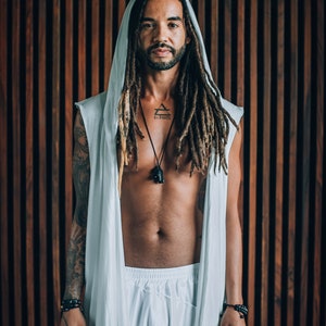 May include: A person with long dreadlocks is wearing a white hooded robe with a sleeveless design. They have a tattoo on their chest that says "Elevate" with a triangle symbol. They are wearing a black necklace with a pendant.