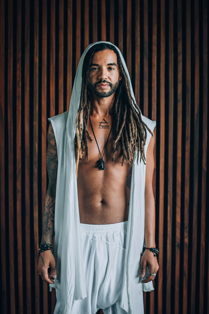 May include: A person with long dreadlocks is wearing a white hooded robe with a sleeveless design. They have a tattoo on their chest that says "Elevate" with a triangle symbol. They are wearing a black necklace with a pendant.