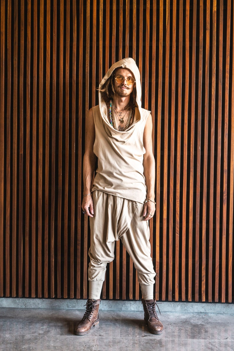 May include: A person wearing a beige hooded top with a draped neckline, beige pants, and brown boots. The person is wearing multiple necklaces and sunglasses. The person is standing in front of a wall of vertical wooden slats.