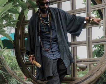 Peut inclure: Une personne portant un chapeau marron, des lunettes de soleil et une veste de style kimono gris foncé. Il est orné de multiples colliers et bracelets. La personne se tient près d'une structure en bambou, avec une flûte en bois à la main.