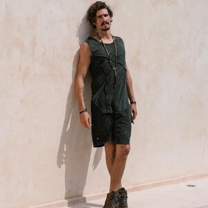 May include: A man wearing a black tank top with a distressed look and black shorts. He is standing against a white wall and wearing a beaded necklace.