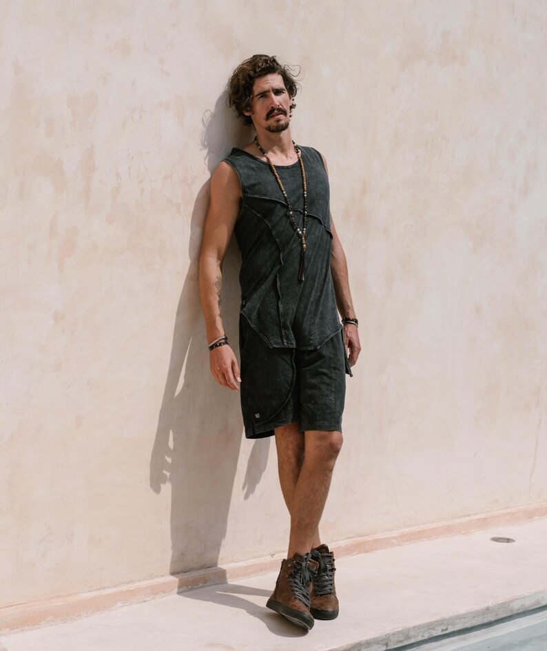 May include: A man wearing a black tank top with a distressed look and black shorts. He is standing against a white wall and wearing a beaded necklace.
